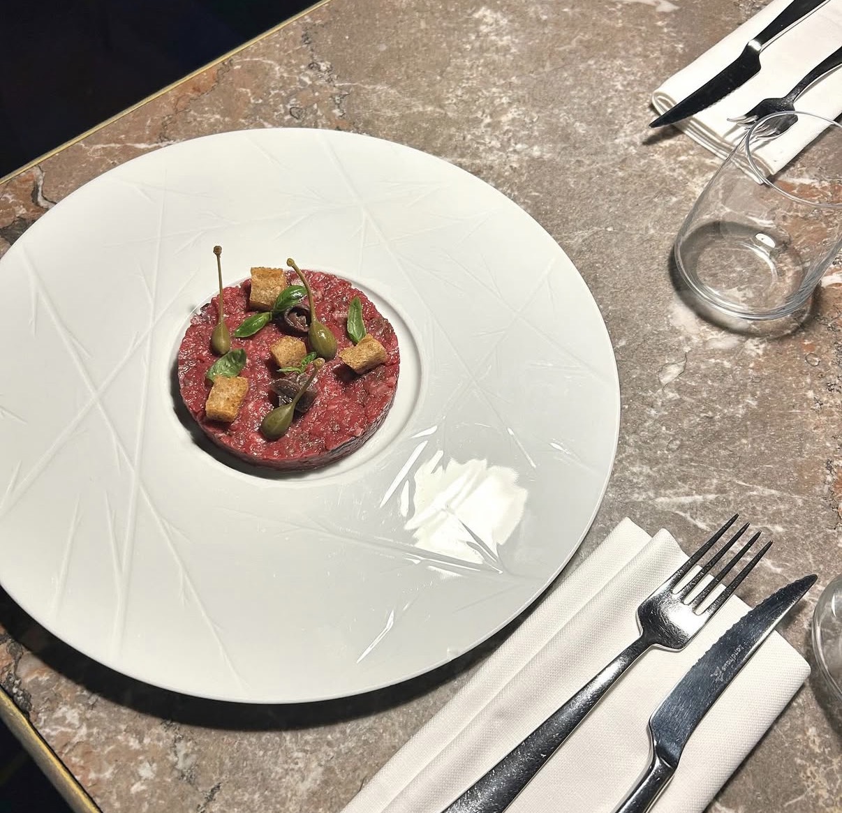Piatto tondo bianco con una preparazione servita su un tavolo in marmo, posate e tovagliolo di lato